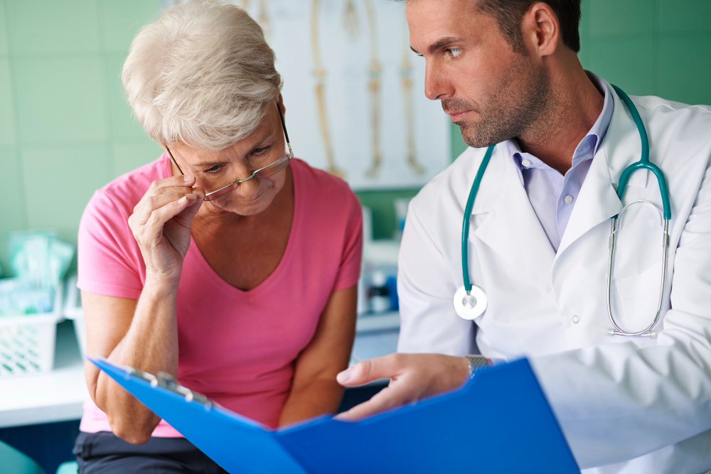 Médico explica informações sobre plano de saúde a uma paciente idosa durante consulta, analisando documentos de cobertura e reajuste.