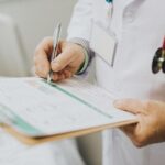 Médico preenchendo um formulário em uma prancheta durante atendimento, com estetoscópio ao fundo, representando consulta médica, prontuário ou documentação clínica.