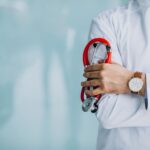 Médico de jaleco branco segurando um estetoscópio vermelho, com fundo azul claro e espaço livre para texto, representando atendimento médico e cuidados de saúde.