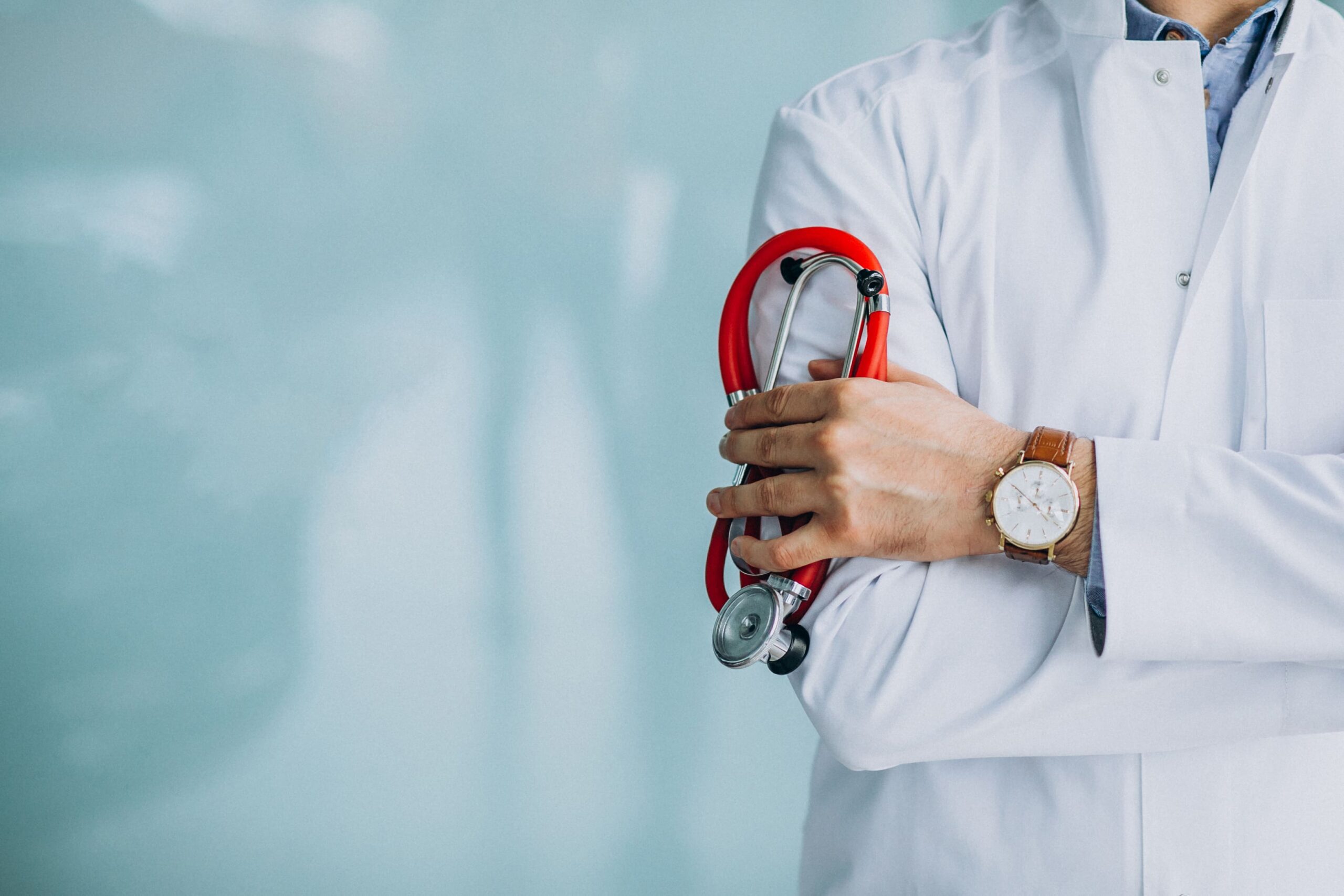 Médico de jaleco branco segurando um estetoscópio vermelho, com fundo azul claro e espaço livre para texto, representando atendimento médico e cuidados de saúde.