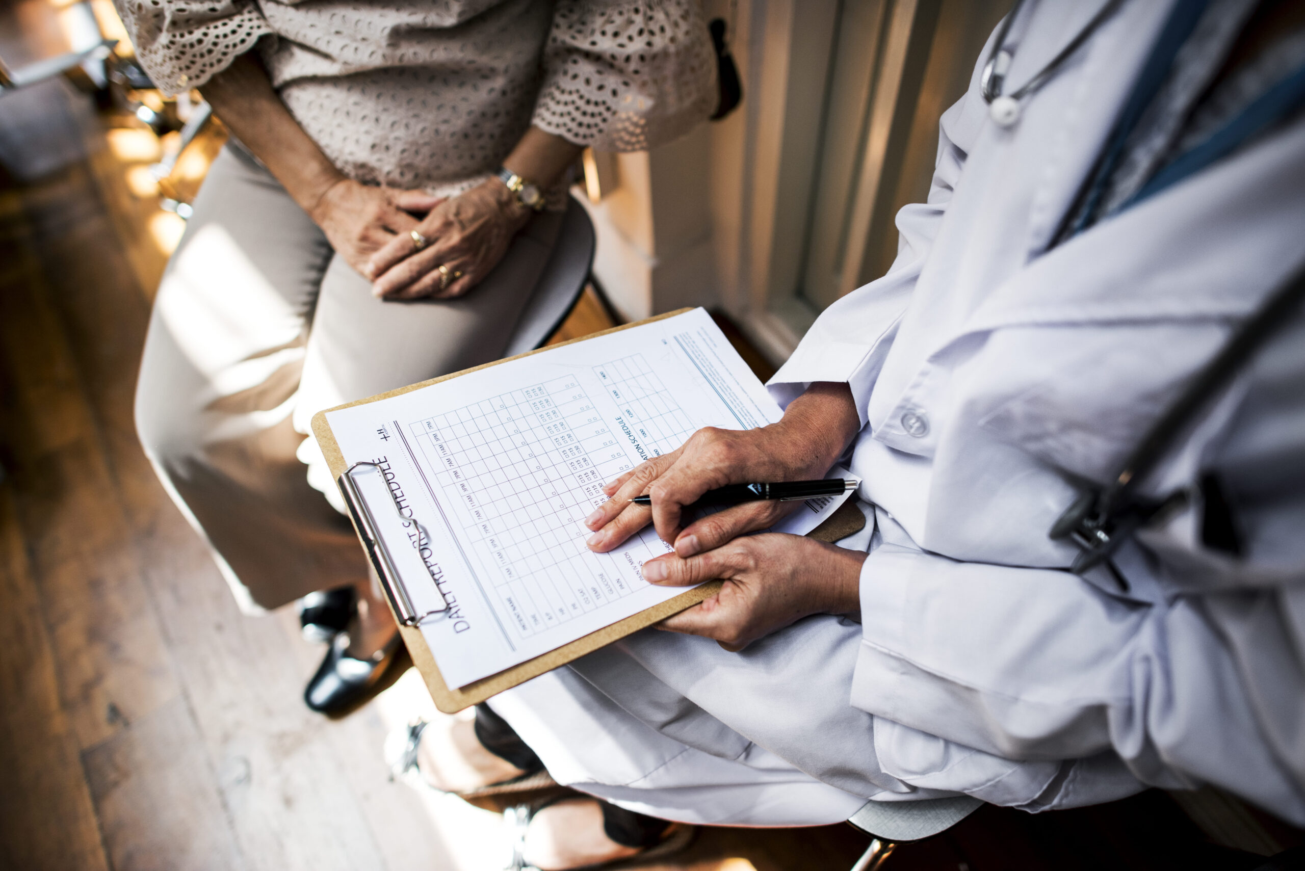 Paciente em consulta médica com profissionais de jaleco, enquanto um médico segura uma prancheta com formulário de agendamento ou prontuário clínico, representando atendimento, avaliação e acompanhamento de saúde.
