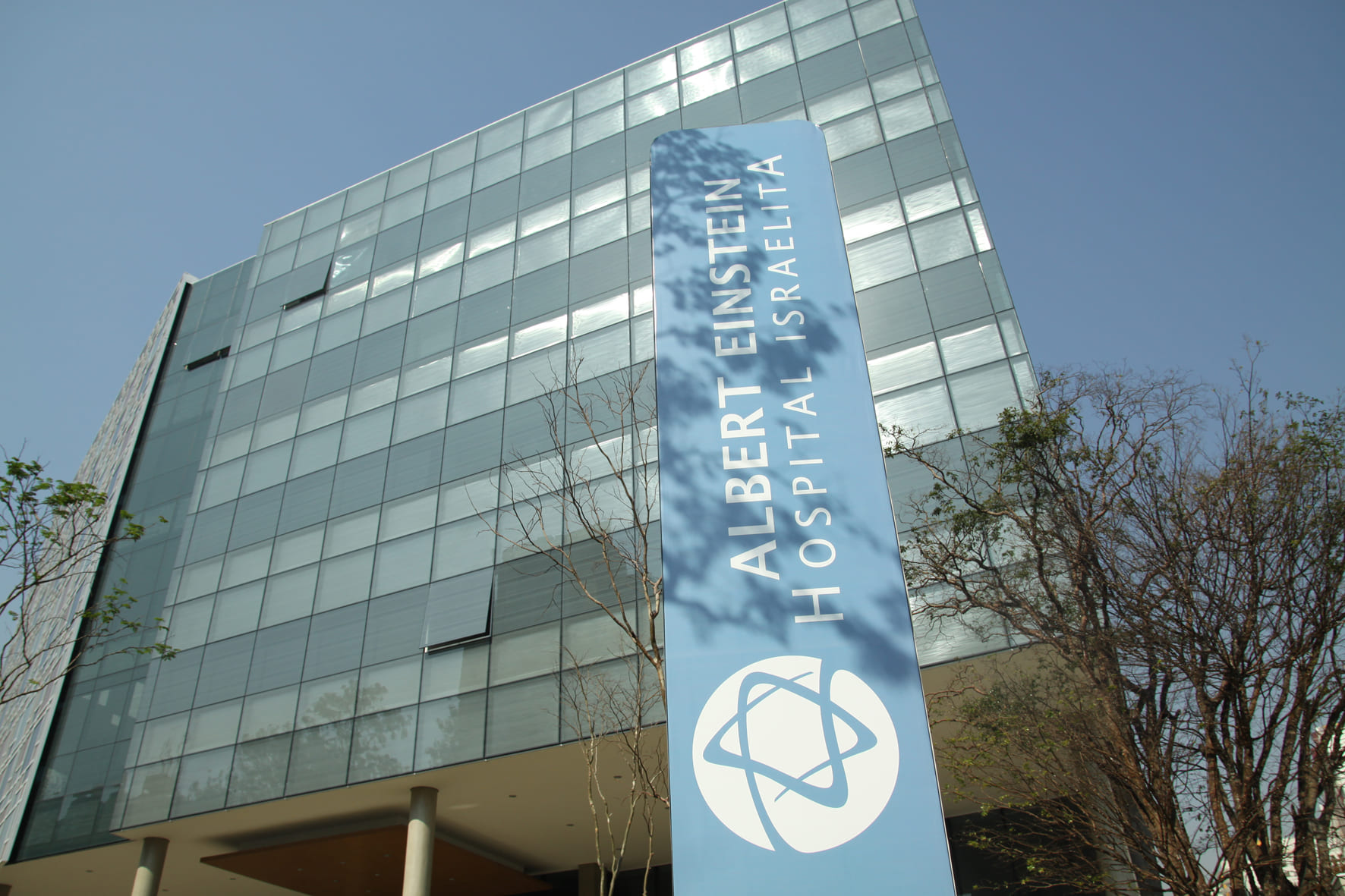 Fachada moderna envidraçada do Hospital Israelita Albert Einstein, com placa azul de identificação em primeiro plano e árvores ao redor, representando uma instituição de saúde de referência.