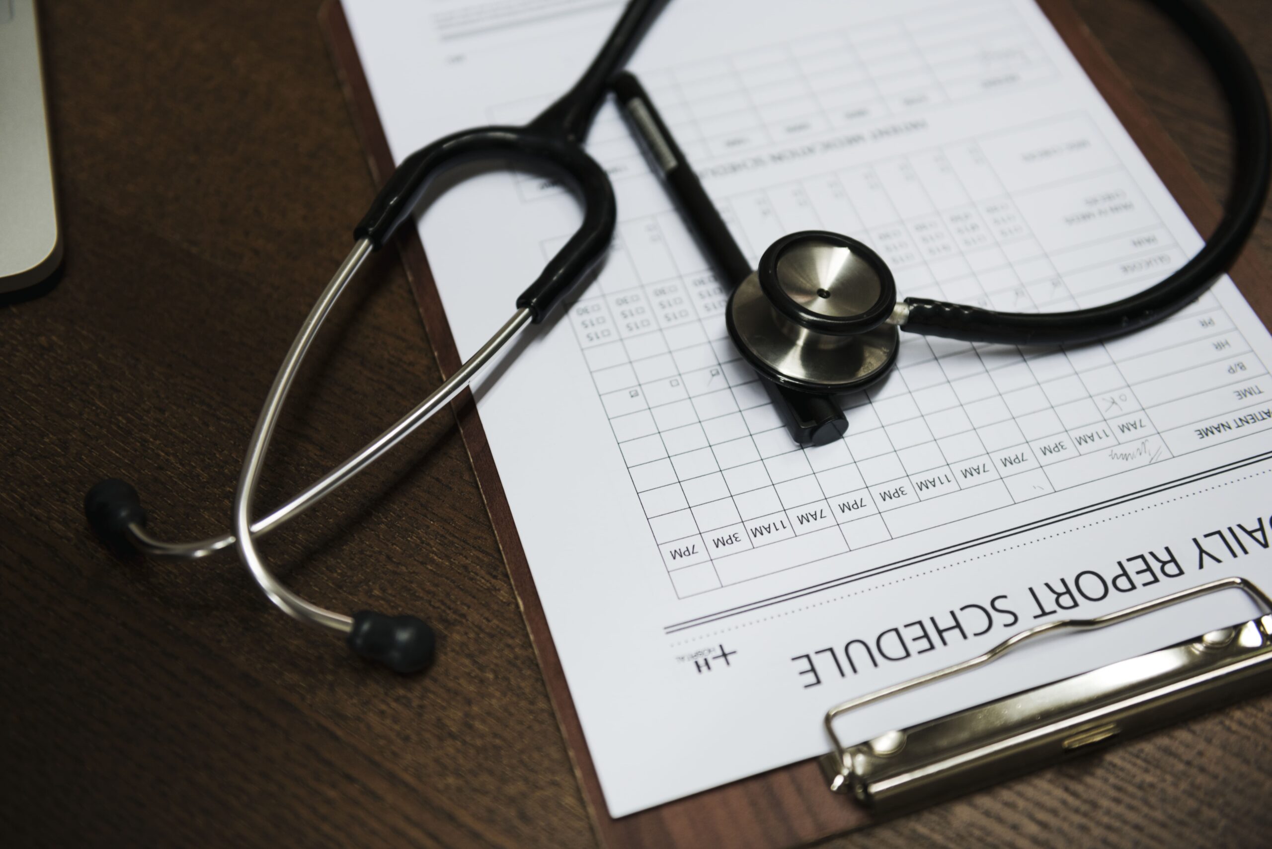 Estetoscópio sobre uma mesa de madeira ao lado de pranchetas com fichas médicas e formulários de agenda de pacientes.